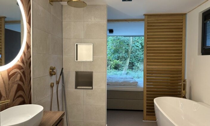 Een moderne badkamer in een Finnhouse Vakantiewoning op Terschelling met ronde spiegel, wastafel links, centraal toilet met inbouwspoeling, vrijstaand bad rechts en houten latten aparte slaapkamer met grote ramen. - Finnhouse