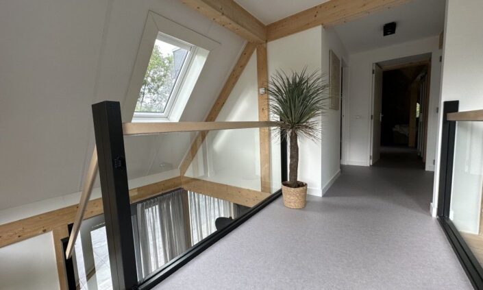 Moderne hal in Finnhouse Vakantiewoning met schuine plafondbalken, dakraam, glazen balustrades. Plantenpot op vloer en deur naar andere kamer de serene charme van Terschelling. - Finnhouse