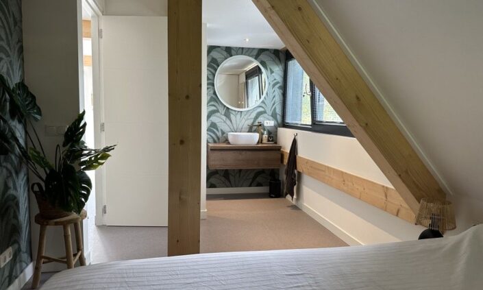 Gezellige slaapkamer in Finnhouse Vakantiewoning Terschelling met zicht op badkamer, houten balken, palmbehang en plant. Badkamer heeft ronde spiegel, houten wastafelmeubel en klein raam. - Finnhouse