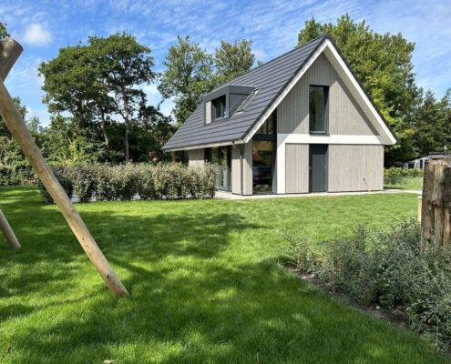 Een modern A-frame huis met donkergrijs dak op een groene grasmat in Finnhouse-stijl. Links staat een houten schommel. Hoge bomen en een keurig gesnoeide haag omringen het terrein. - Finnhouse