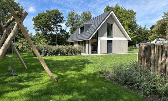 Een modern A-frame huis met donkergrijs dak op een groene grasmat in Finnhouse-stijl. Links staat een houten schommel. Hoge bomen en een keurig gesnoeide haag omringen het terrein. - Finnhouse