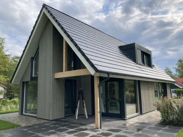 Moderne A-frame Finnhouse met houten gevelbekleding en grote ramen, donker schuin dak met dakkapel. Grijze patio straalt stijl uit, vergelijkbaar met een Vakantiewoning in Terschelling. - Finnhouse