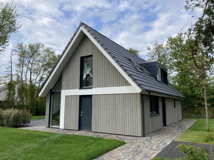 Modern Finnhouse A-frame met lichtgrijze houten gevels en zwart dak, vergelijkbaar met Terschelling vakantiewoning. Grote voorruit en zijdeur, afgesloten door gazon, bestrate paden en bomen onder deels bewolkte hemel. - Finnhouse