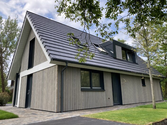 Moderne A-frame Finnhouse met steil, donkergrijs dak en houten gevel. Vakantiewoning verderop door weelderig groen, bomen en gras, onder een deels bewolkte lucht op Terschelling. - Finnhouse