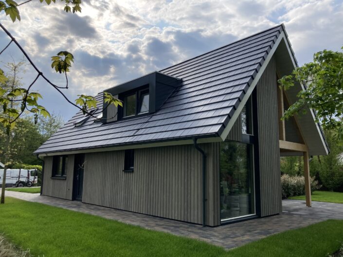 Een eigentijdse houten Finnhouse met schuine, donkere dak en grote ramen, horizontale door groen gras en bomen op Terschelling; de deels bewolkte lucht werpt zacht licht op huis en tuinpad. - Finnhouse