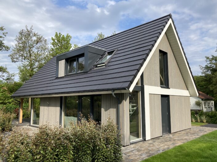Moderne A-frame vakantiewoning met donkergrijs dak, grote ramen en lichte houten gevelbekleding. Omgeven door een netjes aangelegde tuin met struiken en bomen, tegen een lichtbewolkte Terschellingse hemel. - Finnhouse