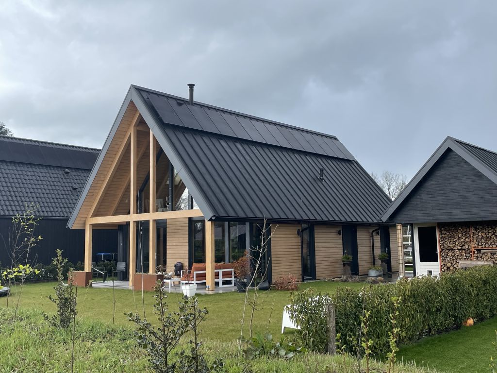 Een moderne A-frame woning met hellend zwart dak en houten gevel, grote glazen ramen en patio zitjes, daarnaast door verzorgde tuin met heg en struiken; weerspiegeling van houtbouw architectuur. - Finnhouse