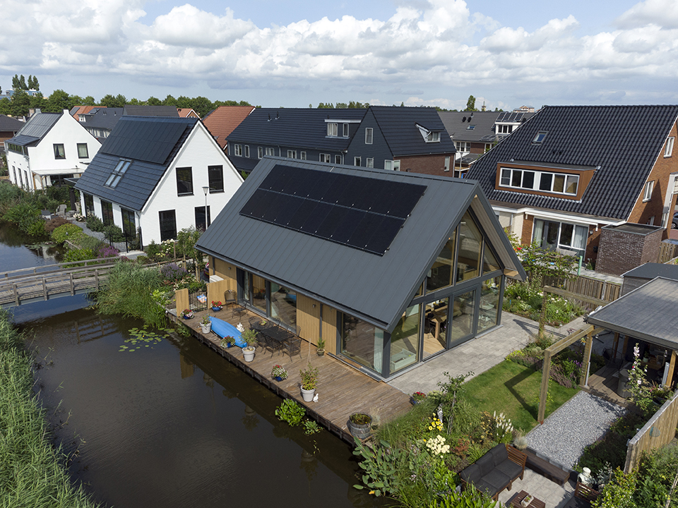 Een moderne Finnhouse met grote glazen ramen en schuin dak met zonnepanelen. Omringd door tuin en water, herhaaldelijk het de essentie van houtbouw naast gelijkwaardige huizen onder een deels bewolkte hemel. - Finnhouse