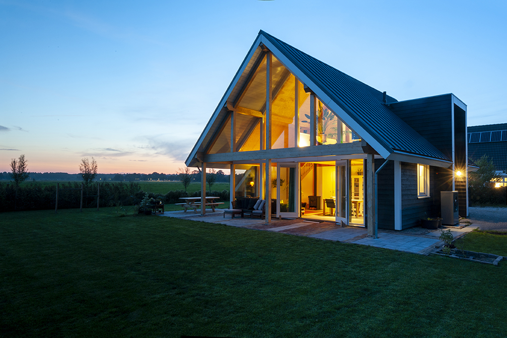 Een moderne houtbouwwoning met grote glazen ramen en een driehoekig dak wordt sfeervol verlicht tijdens het plannen, gelegen op een ruim gazon te midden van een serene natuurlijke omgeving. - Finnhouse