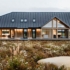 Een moderne Finnhouse in A-frame stijl met grote ramen en donkere metalen daken. Omgeven door wilde bloemen en rotsen, onder een bewolkte lucht temidden van bomen, ideaal voor meergeneratie wonen. - Finnhouse