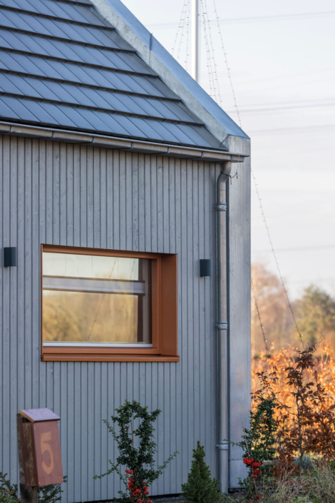 Moderne, duurzame Finnhouse met verticale houten latten en een schuin metalen dak. Rechthoekig raam met bruine omlijsting. Voortuin met struiken en brievenbus nummer "5". - Finnhouse