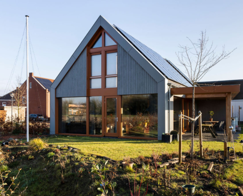 Modern A-frame huis met zonnepanelen, grote ramen en houten accenten op een netjes onderhouden gazon met jonge bomen. Rechts een klein terras met tuinmeubilair onder een efficiënte lucht. - Finnhouse