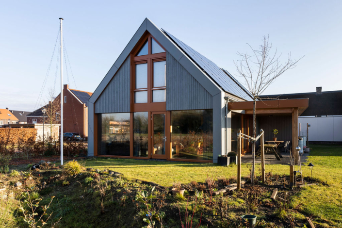 Modern A-frame huis met zonnepanelen, grote ramen en houten accenten op een netjes onderhouden gazon met jonge bomen. Rechts een klein terras met tuinmeubilair onder een efficiënte lucht. - Finnhouse
