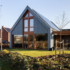 Modern A-frame huis met zonnepanelen, grote ramen en houten accenten op een netjes onderhouden gazon met jonge bomen. Rechts een klein terras met tuinmeubilair onder een efficiënte lucht. - Finnhouse