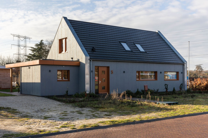 Moderne, grijze Finnhouse met twee horizontaal en schuin dak met dakramen, glas-in-lood voordeur, grote rechthoekige ramen. Omgeven door grindoprit, grasveld en struiken; elektriciteitslijnen op de achtergrond. - Finnhouse