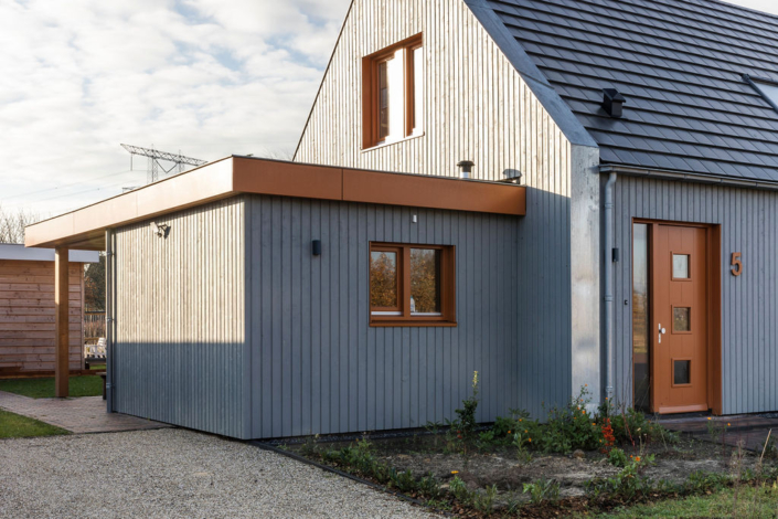 Modern Finnhouse met grijze verticale houten panelen en schuin donker dak, ontworpen volgens duurzame principes. Voorzien van zijuitbouw met luifel, groot voorraam, houten deur nummer 5 en kleine planten langs grindoprit. - Finnhouse