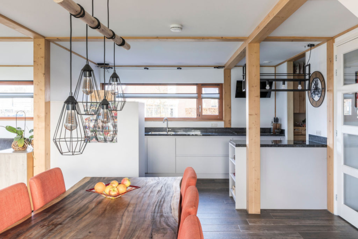 Moderne keuken en eetruimte, houten eettafel met oranje stoelen, appelstuk als centrale, geometrische hanglampen. Duurzaam ontwerp met houten balken, witte kasten en groot raam boven de wastafel. - Finnhouse