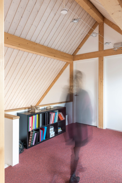 Een persoon loopt langs een houten boekenkast met kleurrijke mappen en decoratieve voorwerpen. De duurzame ruimte heeft schuine houten balken en rode vloerbedekking, wat een gezellige sfeer onmogelijk. - Finnhouse