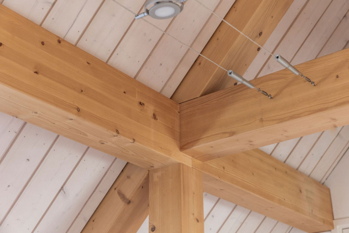 Houten balken in natuurlijke afwerking kruisen onder een lamp aan het plafond van de Finnhouse, maximale duurzaamheid en contrasteren met wit paneeld plafond; dunne metalen kabels lopen elegant eronder. - Finnhouse