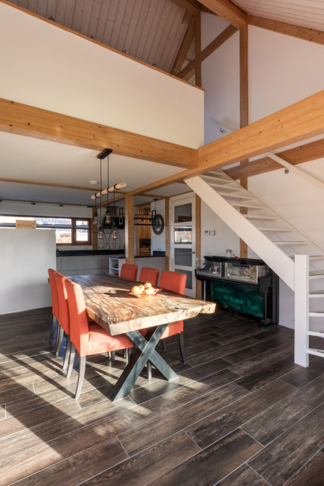 Een eigentijdse eetruimte met duurzame houten tafel, oranje stoelen, donkere houten vloer. Zonlicht door ramen, trap naar loft, zwart omrande glazen kast en minimalistische inrichting creëren een knus geheel. - Finnhouse