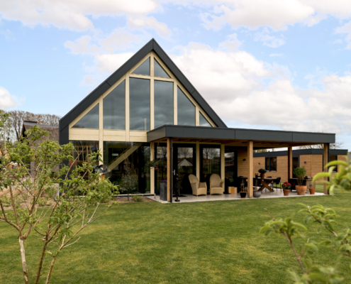 Modern A-frame huis met grote glazen ramen en overdekte patio, omringd door groene tuin, bomen en struiken. Onder deels bewolkte hemel biedt het een duurzame, milieuvriendelijke ontsnapping in de natuur. - Finnhouse