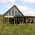 Modern A-frame huis met grote glazen ramen en overdekte patio, omringd door groene tuin, bomen en struiken. Onder deels bewolkte hemel biedt het een duurzame, milieuvriendelijke ontsnapping in de natuur. - Finnhouse