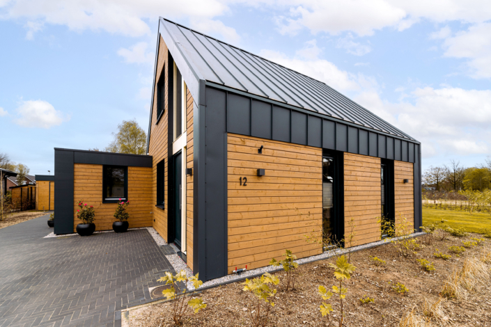 Modern huis met metalen zadeldak en houten gevelbekleding, voorbeeld van duurzaam ontwerp. Oprit van donkere stenen leidt naar entree, horizontale door plantenbakken en jonge beplante omgeving onder bewolkte hemel. - Finnhouse