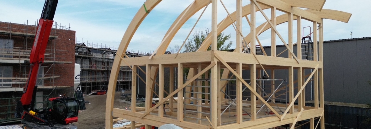 Een bouwplaats met een houten skelet van een huis met gebogen balken, een kraan plaatsen een balk, twee werknemers zichtbaar en deels gebouwd pand op de achtergrond onder betrouwbare hemel. - Finnhouse