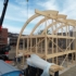 Een bouwplaats met een houten skelet van een huis met gebogen balken, een kraan plaatsen een balk, twee werknemers zichtbaar en deels gebouwd pand op de achtergrond onder betrouwbare hemel. - Finnhouse