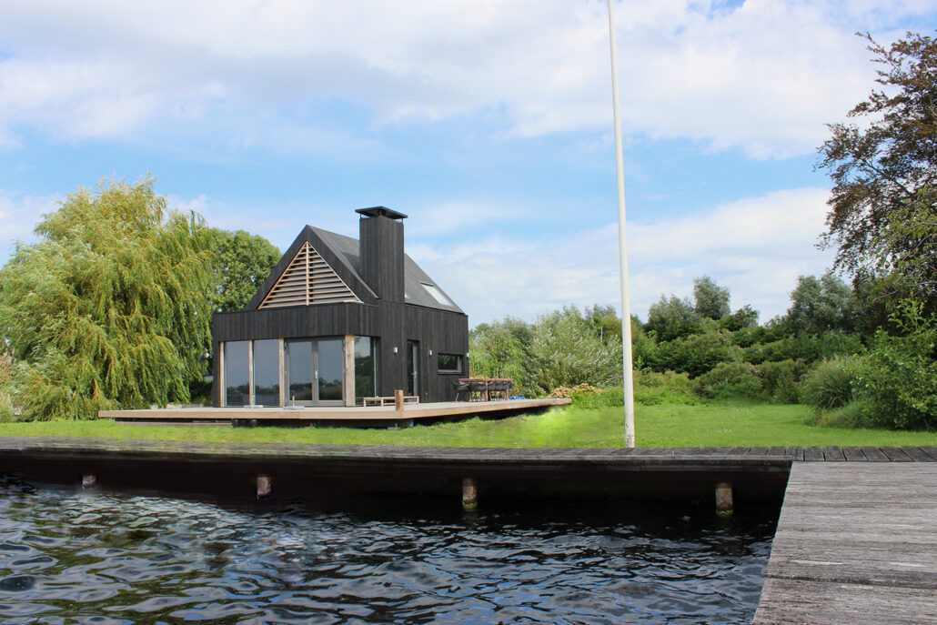 Modern zwart huis met een driehoekig dak en grote ramen bij het water, ideaal voor "eigen huis bouwen," met houten terras en steiger daarbuiten door groenere bomen en bewolkte hemel. - Finnhouse