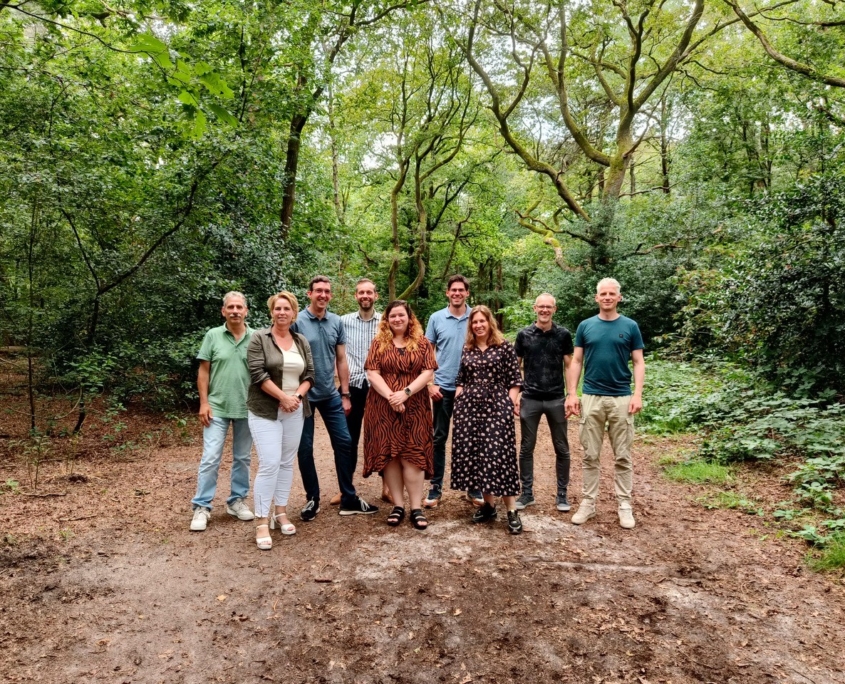 Een groep van tien mensen in een bosrijke omgeving, lachend en informeel gekleed. Groene bomen en bladeren omringen hen, wat schijnbaar op een natuurlijke natuurlijke setting met aarde bedekt. Finnhouse.nl. - Finnhouse
