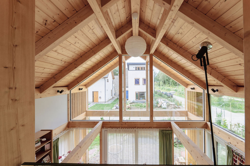 Ruim modern interieur met houten kathedraalplafond en grote driehoekige ramen biedt uitzicht op groene tuin, indirect ecologisch bouwen. Centrale bolvormige hanglamp en microfoonstandaard op de voorgrond zichtbaar. - Finnhouse