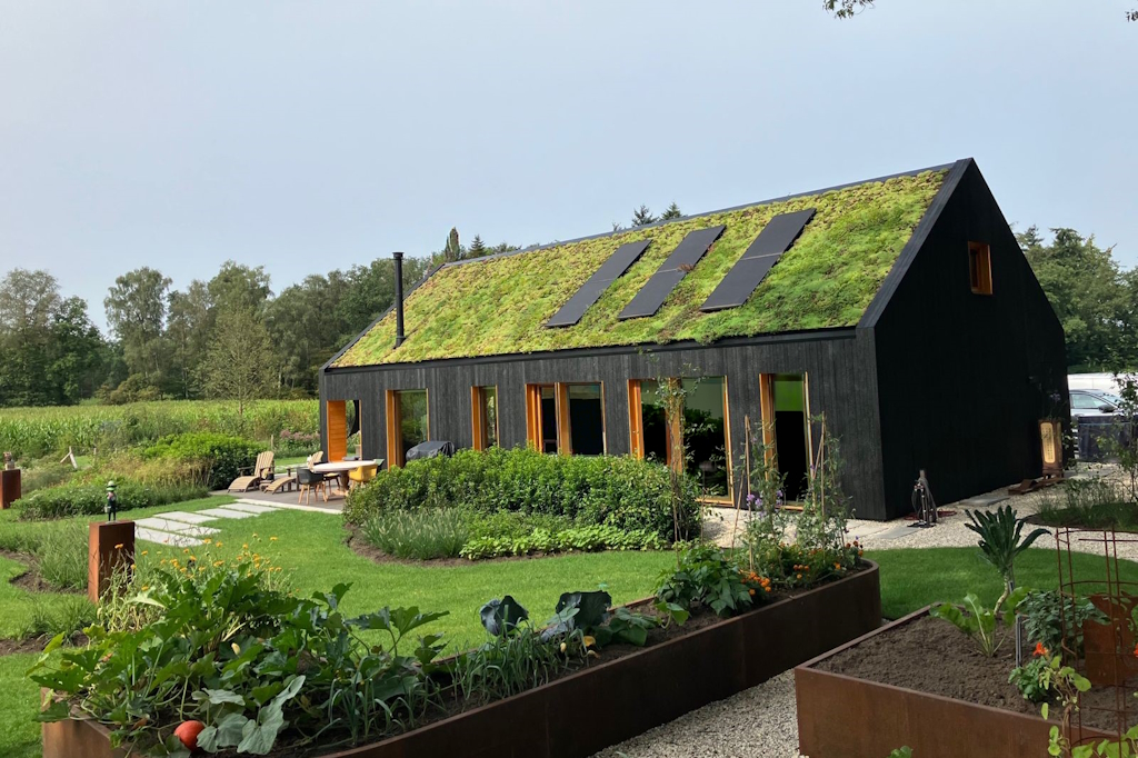 Een modern zwart houten huis met een groen dak is een toonbeeld van ecologisch bouwen. Omgeven door weelderige planten en een tuin verbinden grote ramen het interieur met de natuur, wat bijdraagt aan duurzaam leven. - Finnhouse