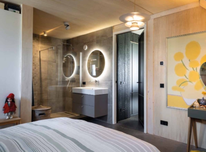 Moderne slaapkamer met en-suite badkamer, inclusief twee ronde spiegels boven een wastafel en een glazen douche. Slaapkamer heeft bed met gestreept beddengoed, pop op kastje en schilderij met groene accenten. - Finnhouse