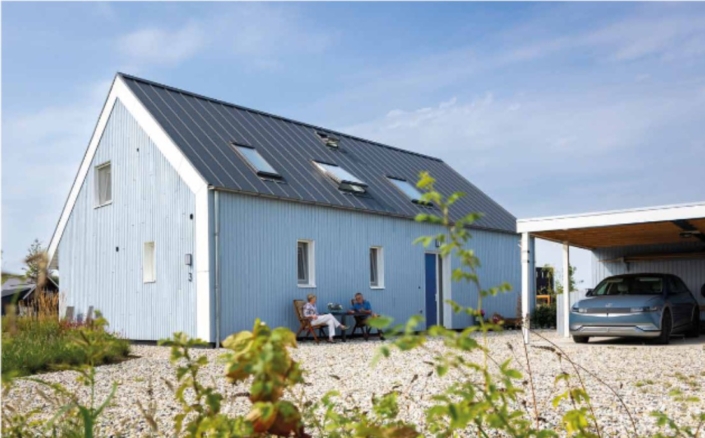 Een modern, blauw huis met een driehoekig dak en dakramen, voorzien van een carport voor een elektrische auto. Twee mensen zitten aan een tafeltje in de voortuin omringd door groene struiken. - Finnhouse