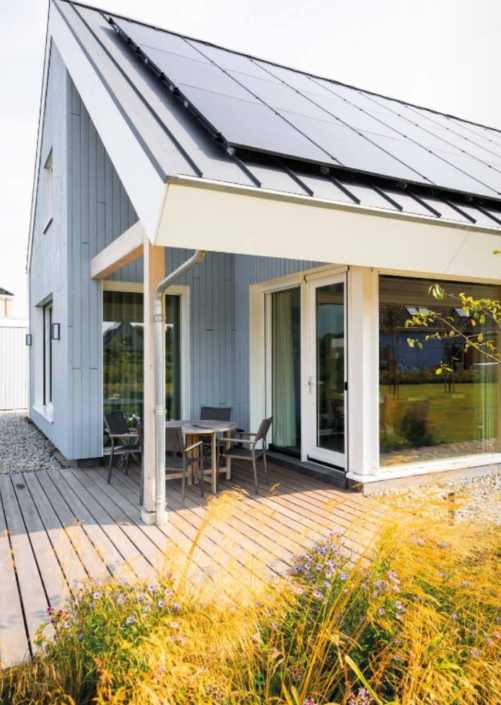 Moderne woning met zonnepanelen op schuin dak en lichtblauwe gevel biedt een duurzame levensstijl. Houten terras met tafel en stoelen te midden van groen, siergrassen en paarse bloemen voor rustgevende sfeer. Finnhouse.nl. - Finnhouse