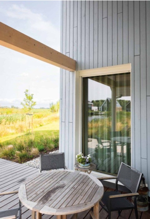 Ronde houten tafel met twee stoelen op terras voor moderne Finnhouse. Grote ramen weerspiegelen groene omgeving met velden en huizen. Bloembak op tafel zorgt voor gezellige sfeer. - Finnhouse