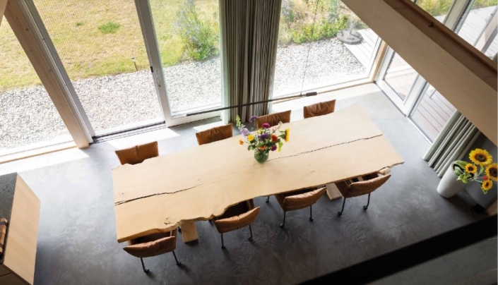 Luchtfoto van een lange houten eettafel met tien stoelen, kleurrijke bloemenarrangement in het midden. Grote ramen en schuifdeur laten natuurlijk licht binnen. Zonnebloemen in vaas, uitzicht op groen gazon. Finnhouse.nl sfeer. - Finnhouse