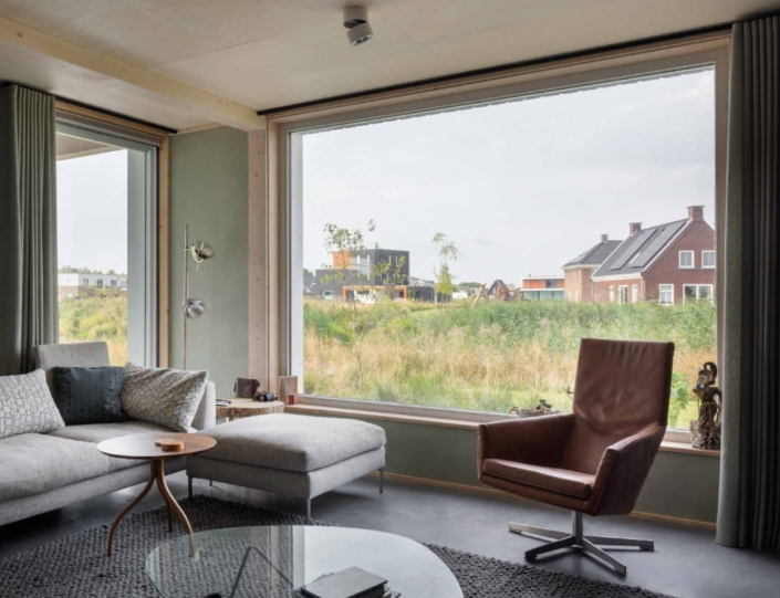 Moderne woonkamer met grote raam biedt uitzicht op groene grasvelden en huizen; binnen een grijze hoekbank met kussens, bruine draaifauteuil, rond houten bijzettafeltje en grijs vloerkleed. Finnhouse.nl. - Finnhouse