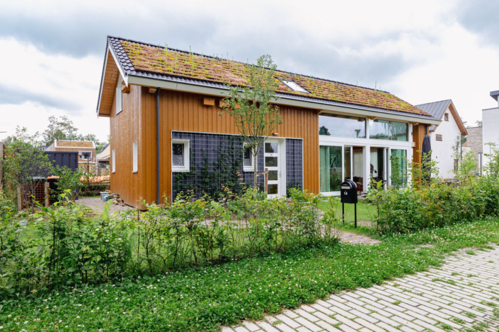 Een modern huis met zadeldak, bedekt met groene mos en planten. Houten bekleding, grote ramen, voortuin met boom en struiken, en een geplaveid pad onder een bewolkte hemel bij Finnhouse.nl. - Finnhouse