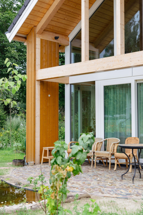 Moderne houten woning met grote glazen ramen en schuin dak, terras met rotan stoelen en tafel. Weelderige tuin met groene flora en een kleine vijver naast het terras. - Finnhouse