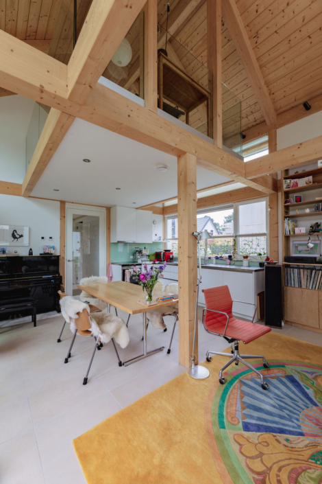 Riant modern interieur met houten balken en hoog plafond. Eethoek heeft schapenvachtstoelen, rode draaistoel en zwarte piano. Keuken met witte kasten, grote ramen en groen vloerkleed. - Finnhouse