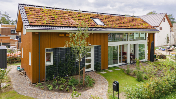 Een moderne, duurzame woning met een groen dak, grote ramen en houten gevel. De tuin heeft een stenen pad, kleine bomen en struiken; op de achtergrond zijn aansluitingen huizen te zien. - Finnhouse