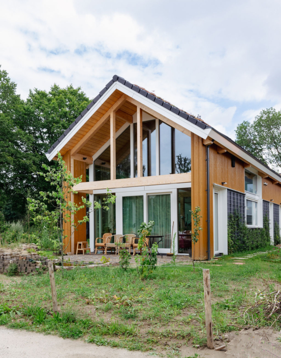 Een modern houten huis met A-frame dak en grote ramen, dalende door gras en groene bomen. Voorzijde heeft een patio met stoelen en tafel, ideaal voor rustige momenten. - Finnhouse