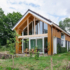 Een modern houten huis met A-frame dak en grote ramen, dalende door gras en groene bomen. Voorzijde heeft een patio met stoelen en tafel, ideaal voor rustige momenten. - Finnhouse