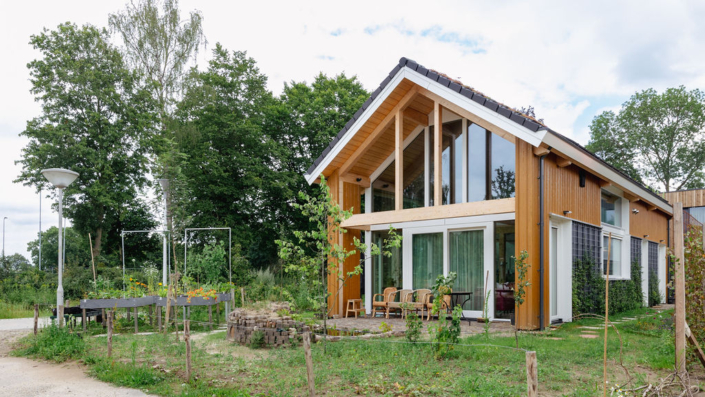 Een modern, houten huis met twee verdiepingen en grote glazen ramen onder een schuin dak schuine deur groen. De voortuin is een groene oase met tafel, stoelen en bomen rond het gazon. - Finnhouse