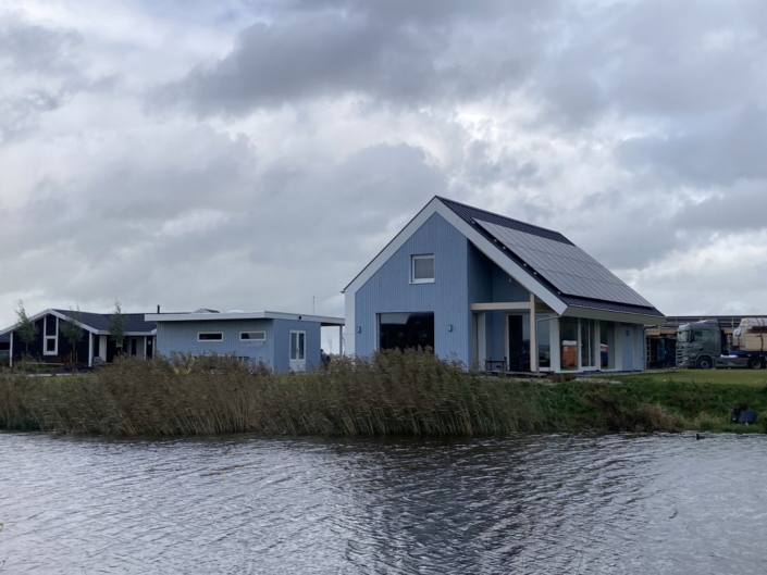 Modern blauw huis met grote ramen en zonnepanelen aan het water, naast een kleiner bouwwerk. Bewolkte lucht en groene zonnige omgeving met wuivend gras langs de waterkant. Finnhouse duurzame architectuur. - Finnhouse
