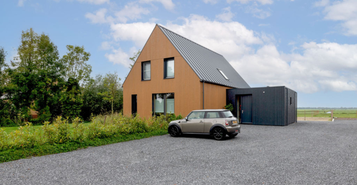 Modern houten huis met twee verdiepingen en schuin metalen dak naast donkere grijze uitbouw; gezinsvriendelijk ontwerp. Grijze auto geïnstalleerd op grindoprit, door gras en bomen onder wolkenlucht. Finnhouse.nl. - Finnhouse