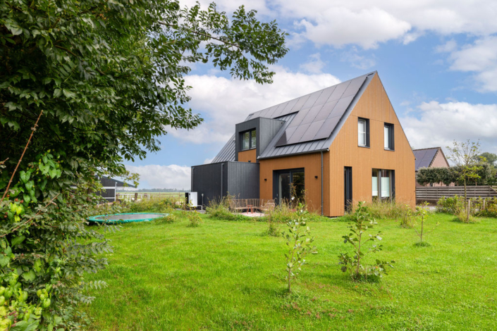 Een moderne, houten woning met twee verdiepingen en donker dak in groene tuin. Omgeven door bomen, planten en trampoline; ideaal familiehuis bij blauwe lucht met wolken. Ideaal voor gezin Finnhouse.nl. - Finnhouse