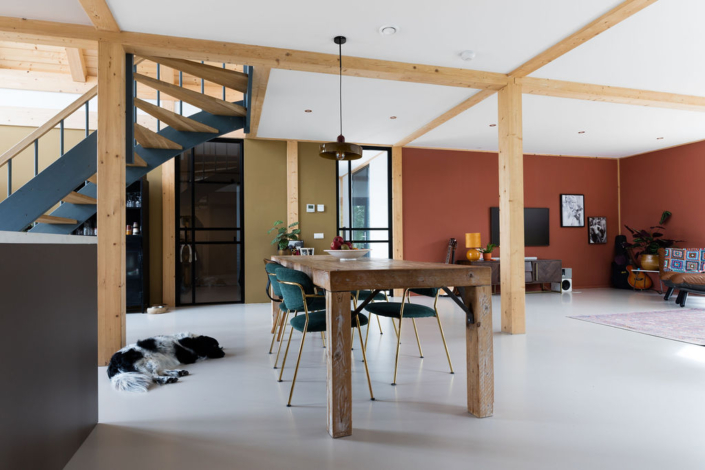 Open leefruimte van Finnhouse met houten eettafel en groene stoelen, ideaal voor gezinsbijeenkomsten. Zwart-witte hond ligt bij trap met zwart hekwerk tussen warme bruine en groene muren. - Finnhouse